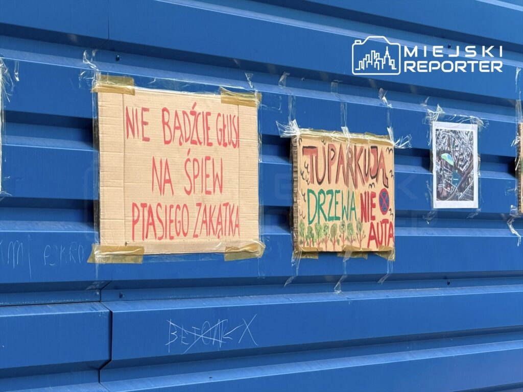 Na niebieskiej ścianie zamocowane są dwa kartony z napisami: "NIE BĄDŹCIE GŁUSI NA ŚPIEW PTAŚCIEGO ZAKATKA" oraz "TUPARKUJA DRZEWA NIE AUTA".
