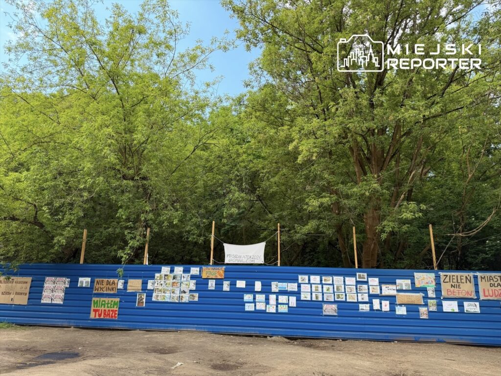 Na niebieskiej ścianie ogrodzenia zawieszone są liczne plakaty i rysunki, w otoczeniu zielonych drzew.