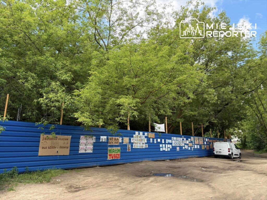 Na niebieskiej ogrodzeniu umieszczone są różnorodne plakaty i rysunki, obok stoi biały dostawczy samochód, w tle gęste drzewa.