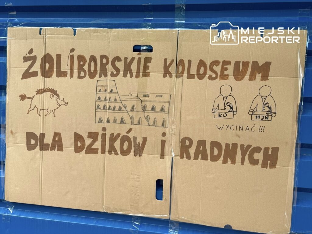 Kartonowa plansza z napisem "Żoliborskie Koloseum dla dzików i radnych", z rysunkiem dzika oraz postaciami przy mównicach.