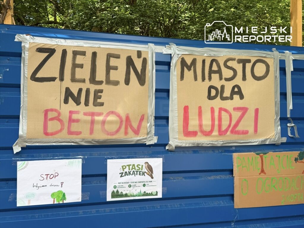 Na niebieskiej ścianie umieszczono kartonowe plakaty z hasłami: "ZIELEN' NIE BETON", "MIASTO DLA LUDZI" oraz rysunek z napisem "STOP Wycinanie drzew".