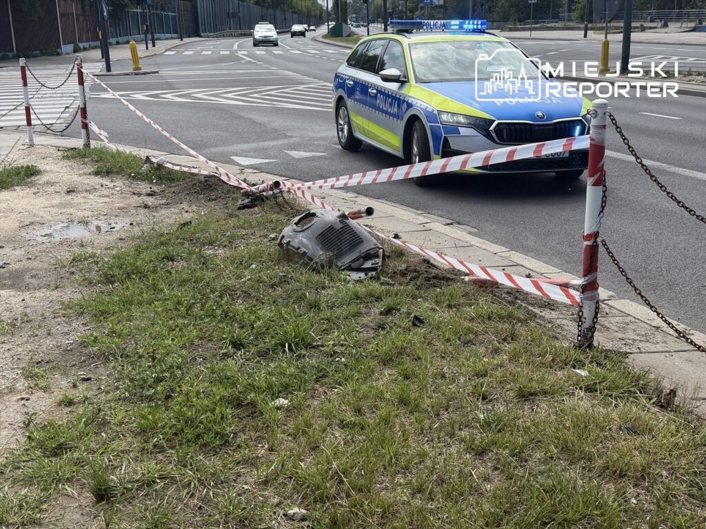 Policjant w radiowozie z włączonymi sygnałami świetlnymi monitoruje miejsce zdarzenia, zabezpieczone taśmą.