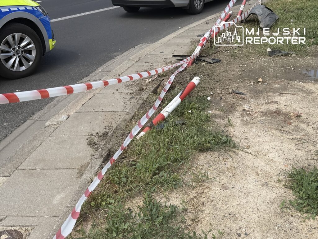 Na chodniku widoczne są czerwono-białe taśmy zabezpieczające, obok leży uszkodzone auto, a w tle przejeżdża inny pojazd.