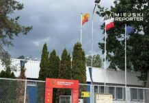Wejście do akademii piłkarskiej z flagami Polski, Unii Europejskiej i regionu, otoczone wysokimi tujami.