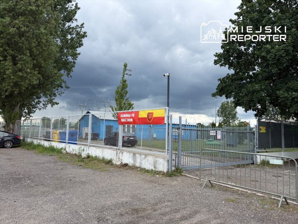 Bramka wejściowa na teren obiektu sportowego z niebieskim budynkiem w tle i flagą z napisem "FLAMINGI TO NASZ DOM".