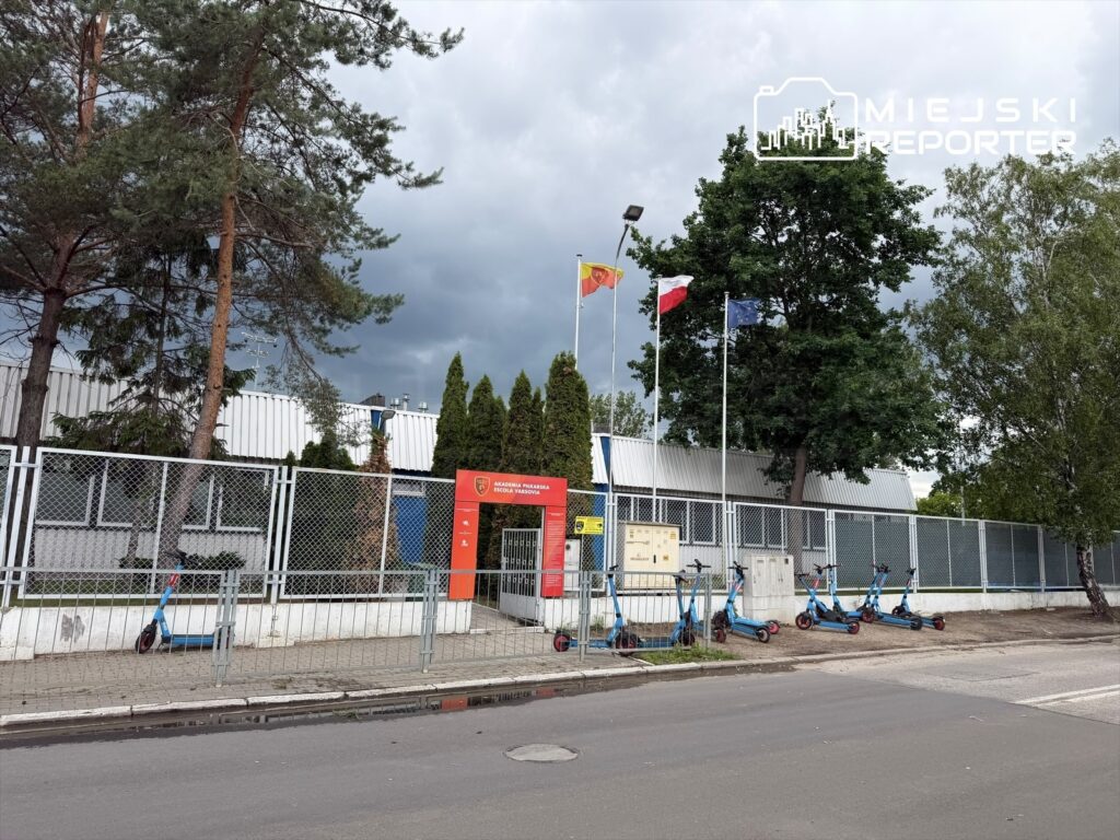 Budynek Akademii Przyszłości z flagami na maszcie, w tle ciemne chmury, a przed nim stoją niebieskie hulajnogi.