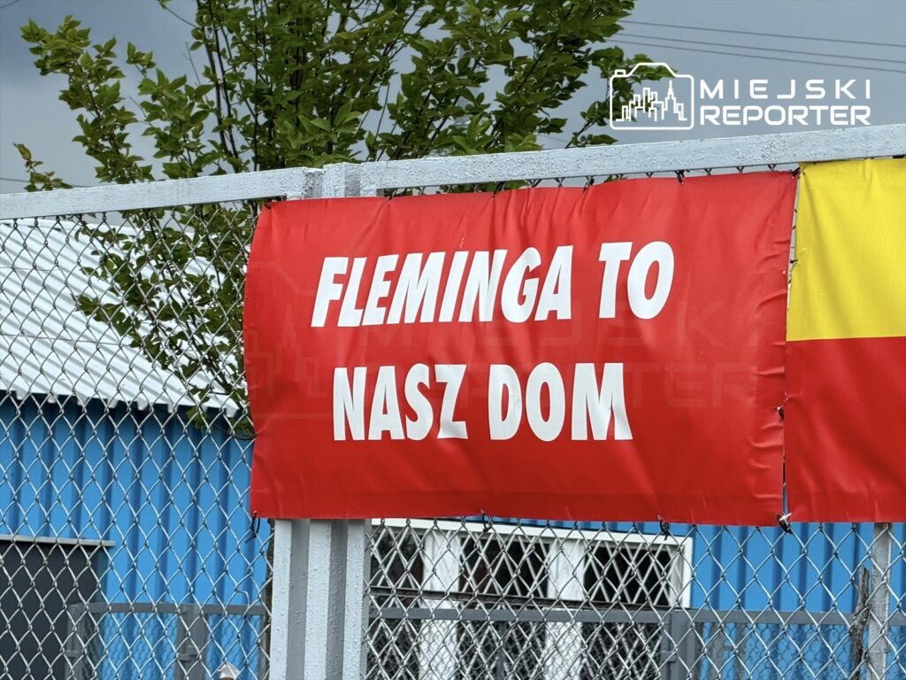 Czerwony baner z napisem "FLEMINGA TO NASZ DOM" zawieszony na ogrodzeniu, w tle niebieski budynek.