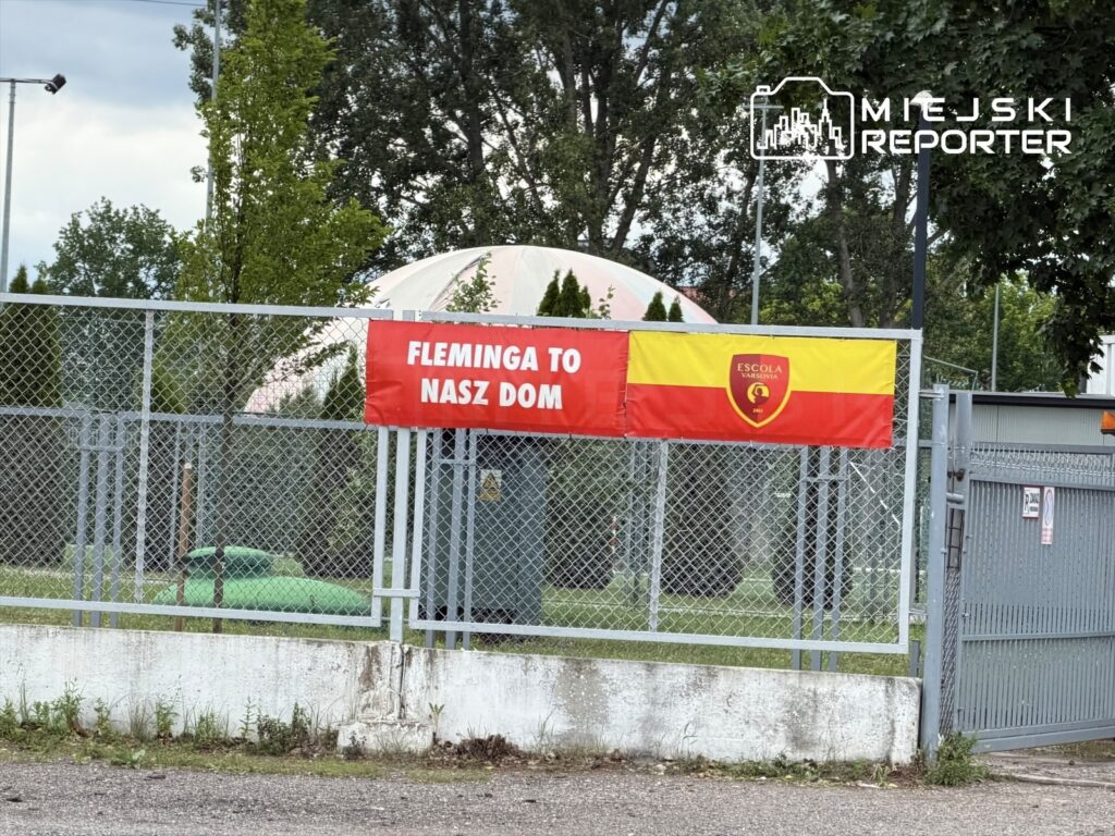 Na ogrodzeniu obiektu sportowego powiewają flagi z hasłem "Fleminga to nasz dom" oraz logo Escola Varsovia.