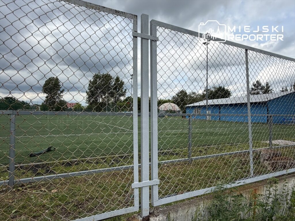 Zamknięta brama na boisku sportowym z sztuczną nawierzchnią, w tle niebieski budynek i chmurne niebo.