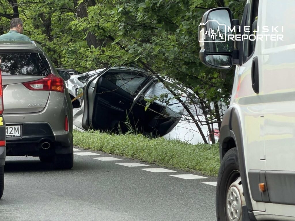 Na drodze leży przewrócone auto, a obok stoi mężczyzna, obserwujący sytuację. W tle widać inne pojazdy.