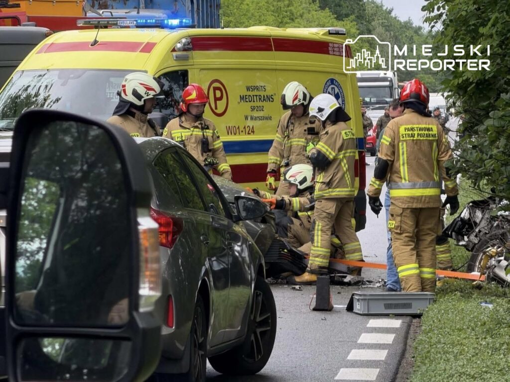 Grupa strażaków w mundurach przystępuje do akcji ratunkowej obok rozbitego pojazdu na drodze.