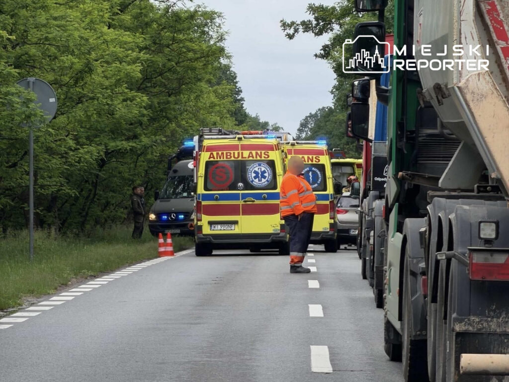 Karetki pogotowia oraz pojazdy służb ratunkowych zatrzymane na drodze, obok mężczyzna w pomarańczowej kamizelce.