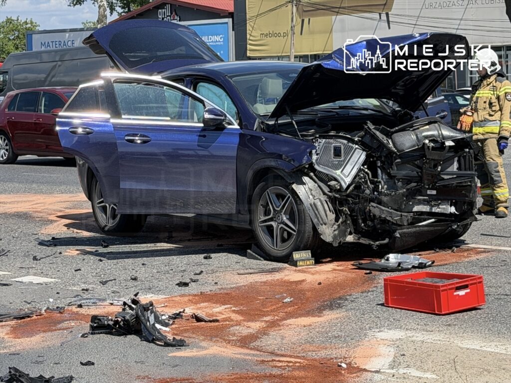 Niebieski samochód osobowy z uszkodzoną przednią częścią stoi na drodze, obok niego leżą fragmenty pojazdu i plama cieczy.