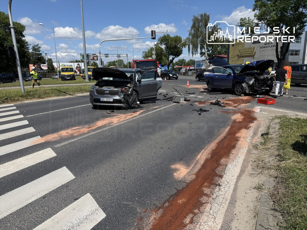 Dwa rozbite samochody na skrzyżowaniu, wokół leżą części pojazdów i plamy na asfalcie. W tle widać inne pojazdy i sygnalizację świetlną.