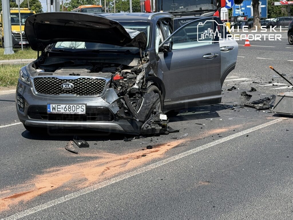 Uszkodzony samochód osobowy marki Kia z otwartą maską na jezdni, wokół widać pozostałości po wypadku i ślady na asfalcie.