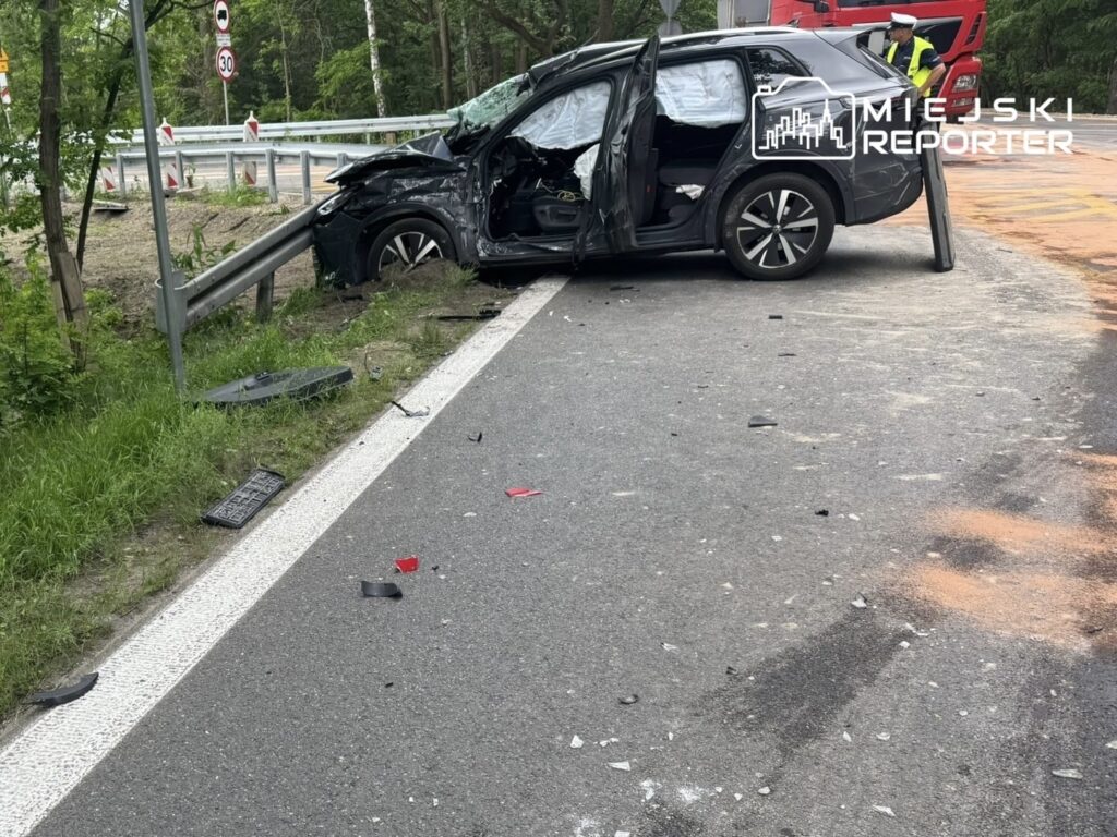Czarny samochód osobowy z otwartymi drzwiami stoi na poboczu drogi, otoczony odłamkami i uszkodzeniami po wypadku.