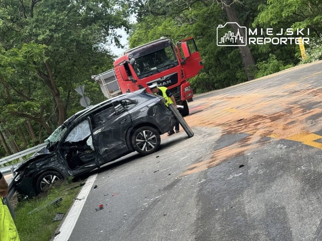 Czarny samochód osobowy z uszkodzoną stroną stoi na drodze, obok niego czerwony ciężarowy pojazd oraz ślady płynów na asfalcie.