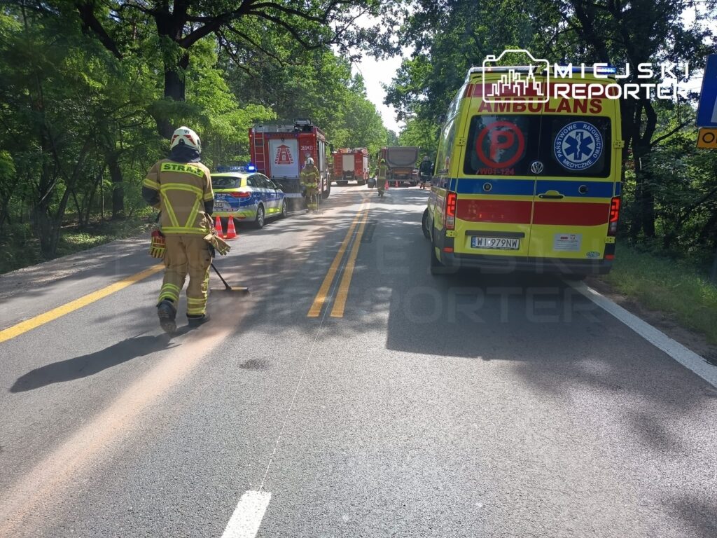 Strażak w stroju ochronnym sprząta drogę, obok stoją pojazdy służb ratunkowych w lesistym otoczeniu.