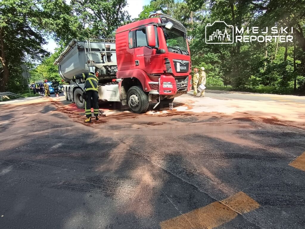Czerwony ciężarówka MAN z rozlaną substancją na drodze, obok strażacy w akcji w leśnym otoczeniu.