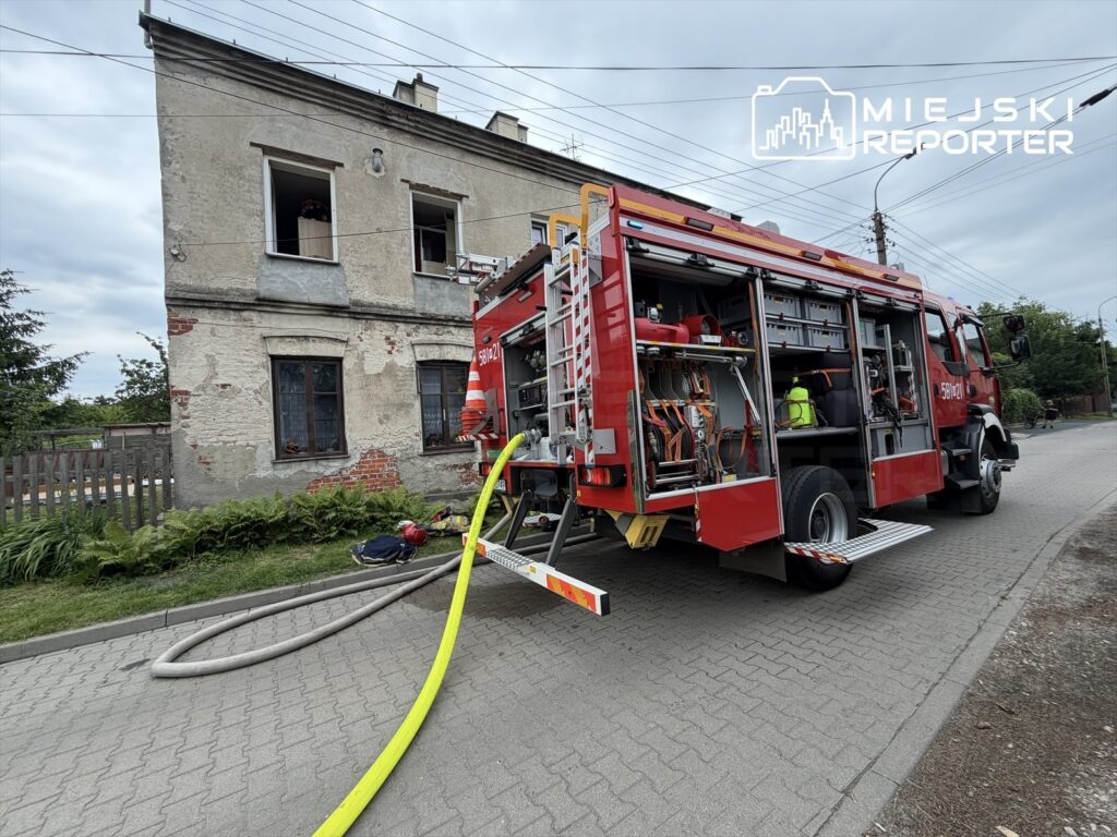 Wóz strażacki z rozwiniętym wężem stoi przed budynkiem mieszkalnym z uszkodzonymi oknami.