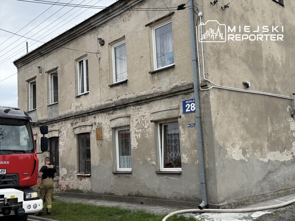 Strażak przy czerwonym wozie strażackim sprawdza budynek mieszkalny z widocznymi śladami zużycia.