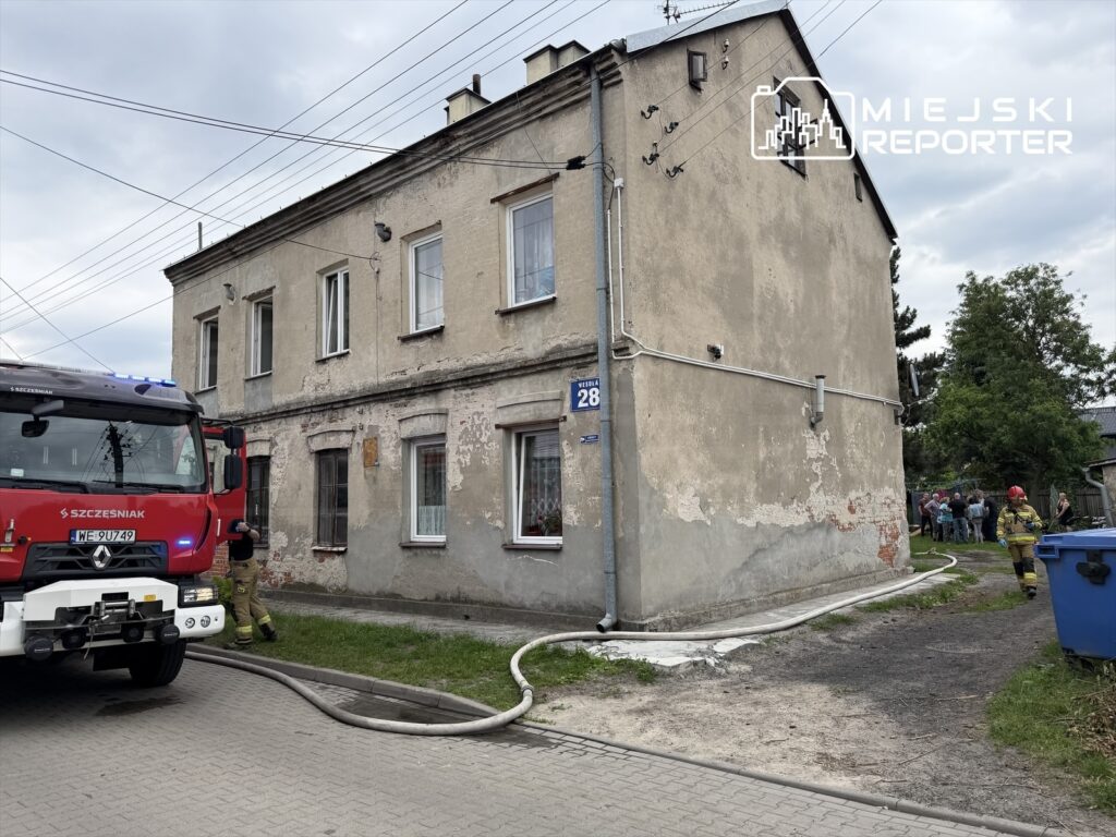 Woz z strażacki zatrzymany przy budynku mieszkalnym, obok grupy obserwatorów, w tle widoczna uszkodzona elewacja.