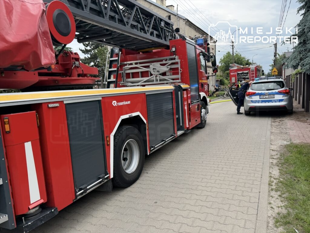 Wóz strażacki z podnośnikiem zaparkowany obok radiowozu policyjnego na ulicy, obok budynków mieszkalnych.