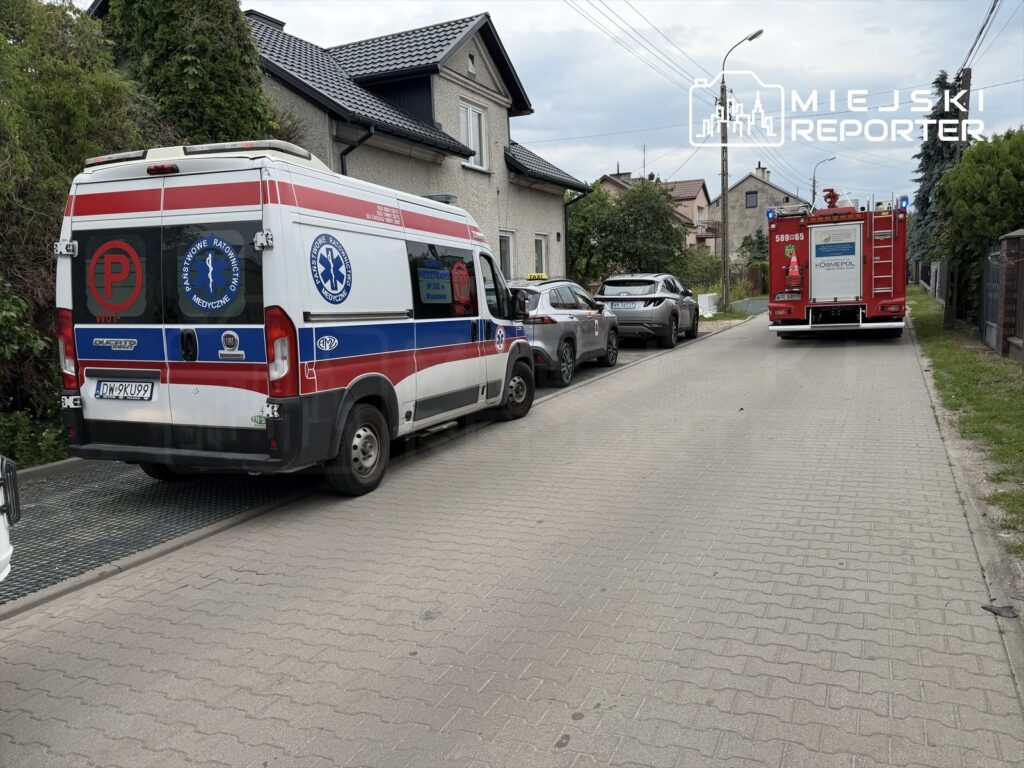 Karetka pogotowia i wóz strażacki zaparkowane na wąskiej ulicy osiedlowej, otoczone samochodami osobowymi.