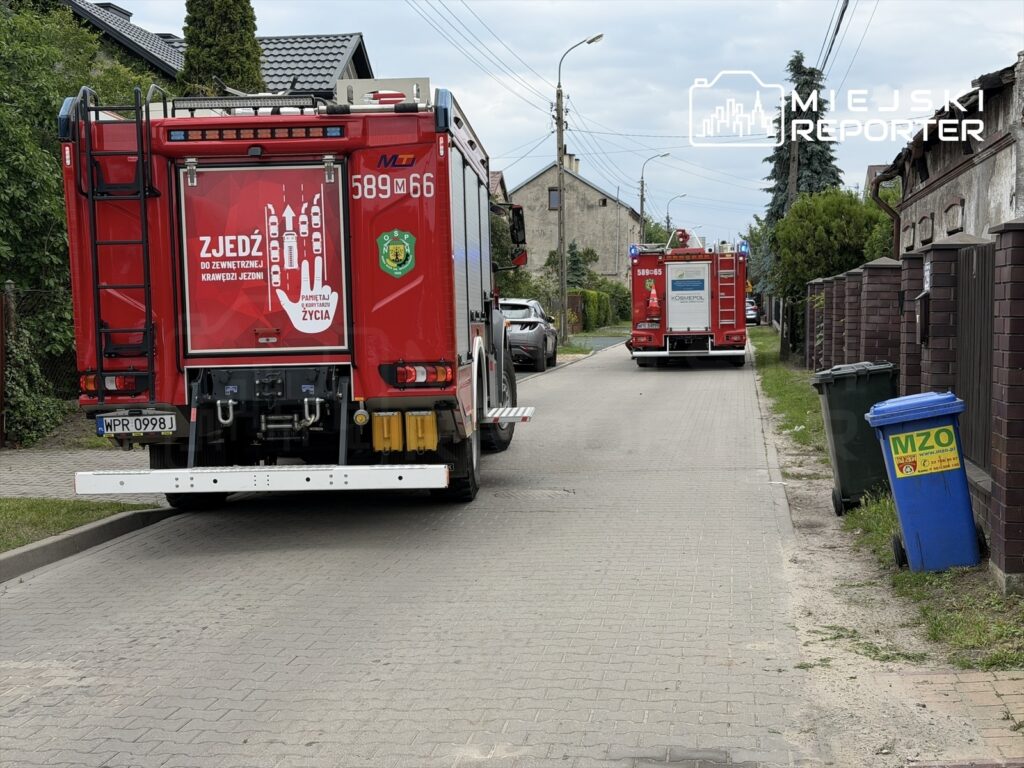 Dwa wozy strażackie stoją na wąskiej ulicy, obok nich znajduje się niebieski pojemnik na odpady z napisem "MZO".