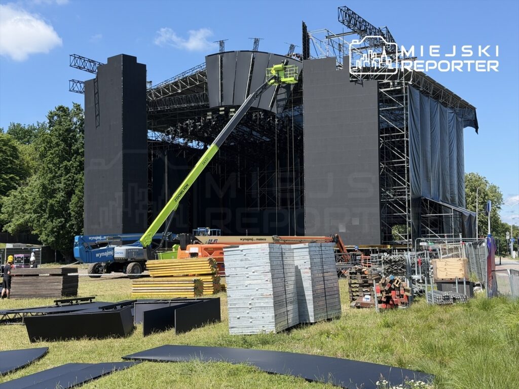 Ekipa techniczna montuje scenę festiwalową w terenie zielonym, obok stoją sprzęty i materiały budowlane.