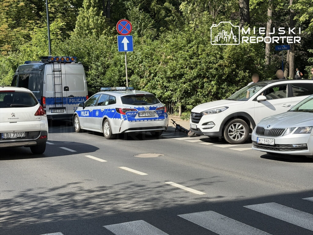 Policyjny radiowóz oraz furgonetka zatrzymane na ulicy, obok zaparkowane samochody i znak zakazu zatrzymywania się.