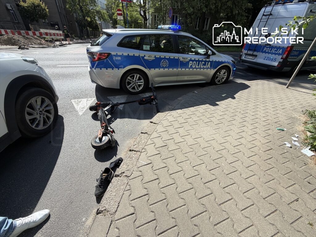 Policyjny radiowóz z włączonymi sygnałami świetlnymi blokuje ulicę obok przewróconej hulajnogi i porzuconego obuwia.