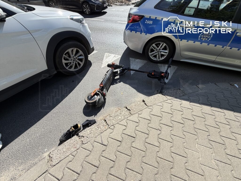 Na drodze leży uszkodzona hulajnoga elektryczna obok zaparkowanych samochodów, obok niej znajduje się jeden but i czarna odzież.