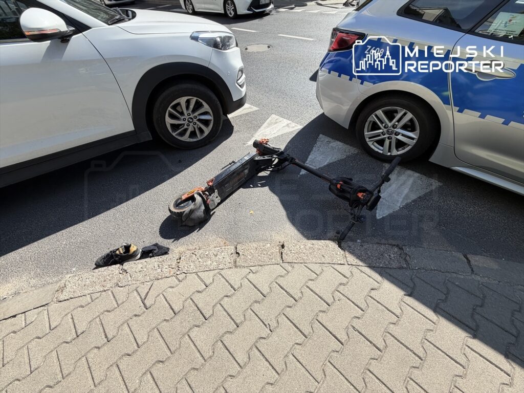 Na chodniku leży uszkodzona hulajnoga elektryczna obok dwóch zaparkowanych samochodów, a obok widać skarpetę i but.