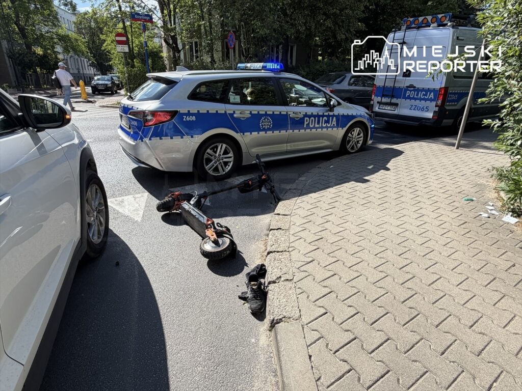 Policyjny radiowóz z włączonymi sygnałami świetlnymi stoi na skrzyżowaniu obok przewróconej hulajnogi elektrycznej.