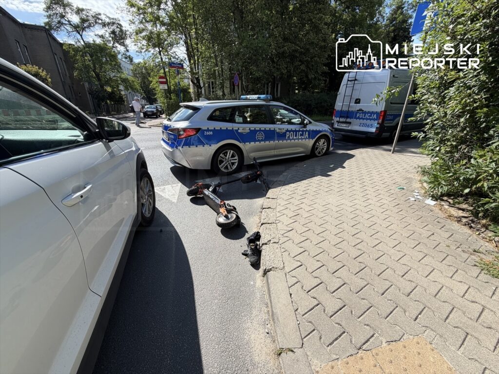Na jezdni leżą dwa uszkodzone hulajnogi elektryczne, obok nich zaparkowany radiowóz policyjny. W tle widać zieleń drzew.