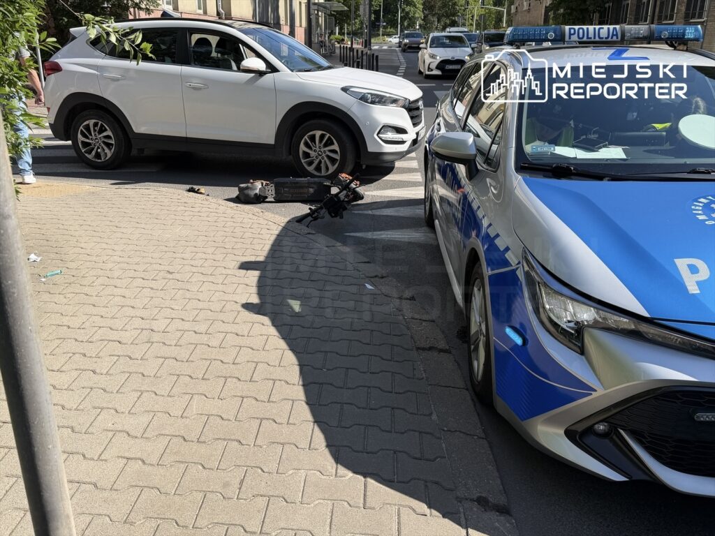 Biały samochód osobowy zatrzymany na drodze obok policyjnego auta, na chodniku leży uszkodzone wyposażenie.