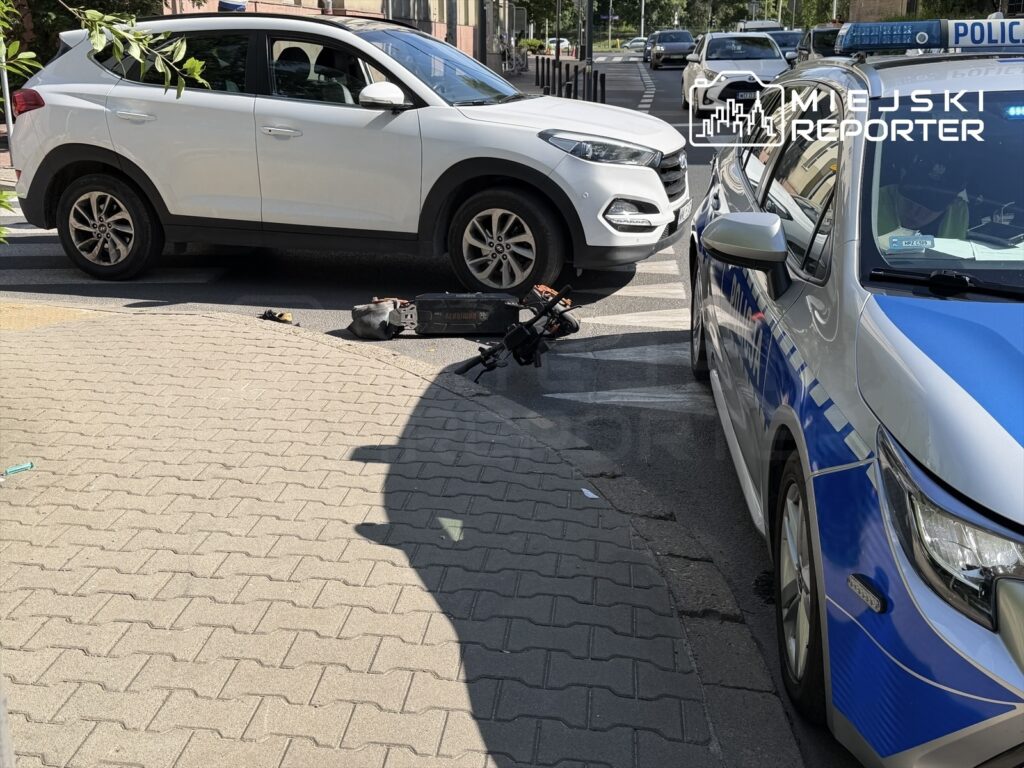 Biały samochód osobowy zablokowany na skrzyżowaniu obok policyjnego radiowozu, na jezdni leży uszkodzony skuter.