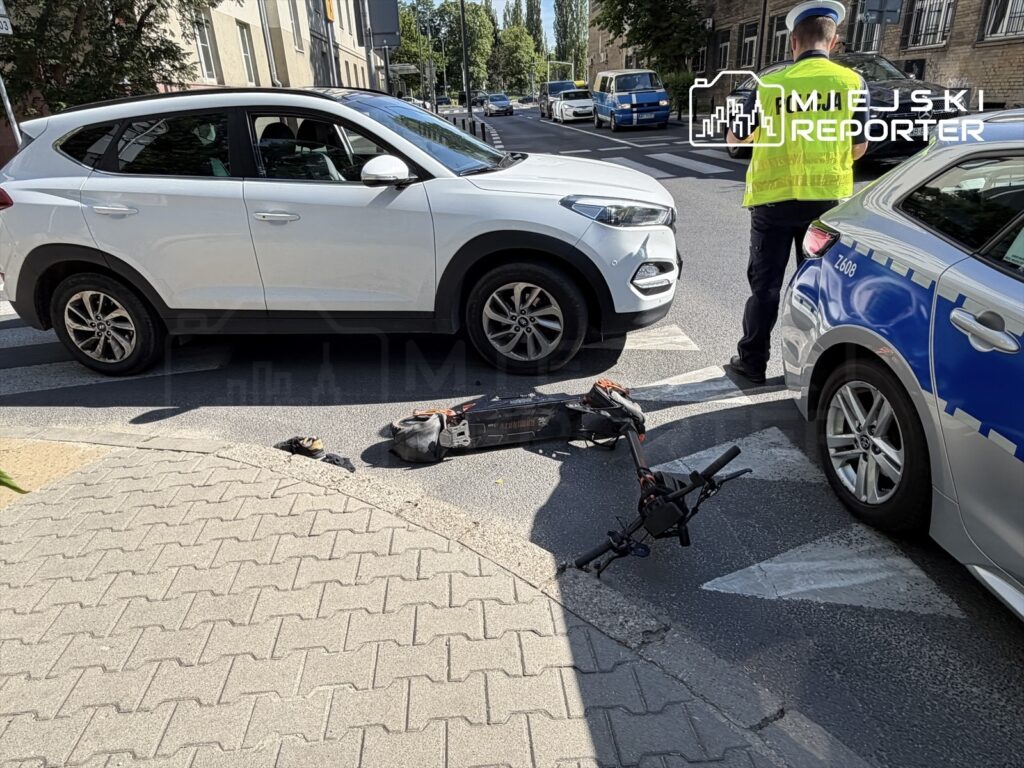 Na jezdni leży uszkodzony skuter elektryczny, obok zatrzymane białe auto osobowe oraz radiowóz policyjny, przy którym stoi policjant.