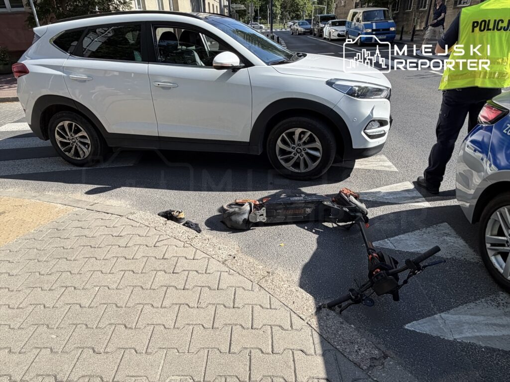 Na jezdni leży uszkodzony skuter elektryczny, obok zatrzymany biały samochód osobowy; w tle policjant rozmawia z kierowcą.