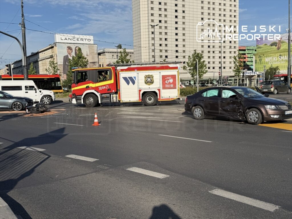 Na skrzyżowaniu stoi wóz strażacki, a obok leży uszkodzony motocykl oraz zderzona ciemna osobówka z wgniecioną stroną.