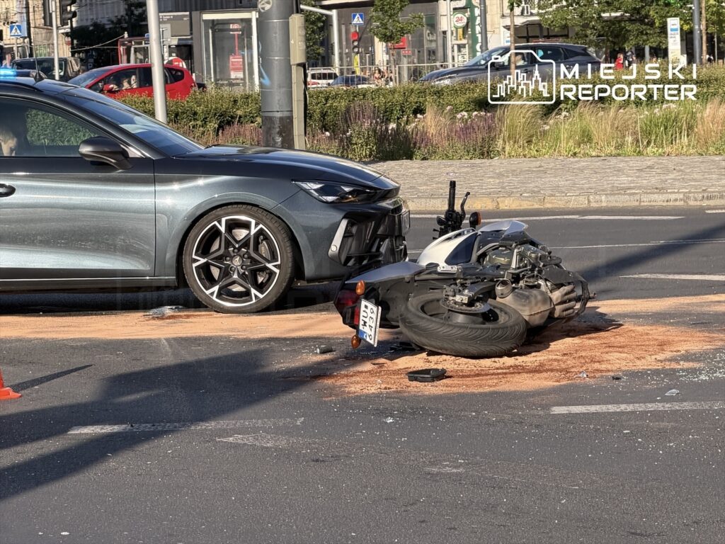 Motocykl leży na jezdni obok uszkodzonego samochodu, a na drodze widoczny jest rozlany płyn.