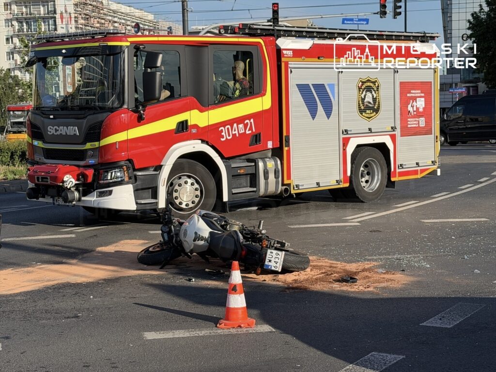 Strażacki wóz Scania zatrzymany na skrzyżowaniu obok przewróconego motocykla na drodze, obok leży plama płynu.