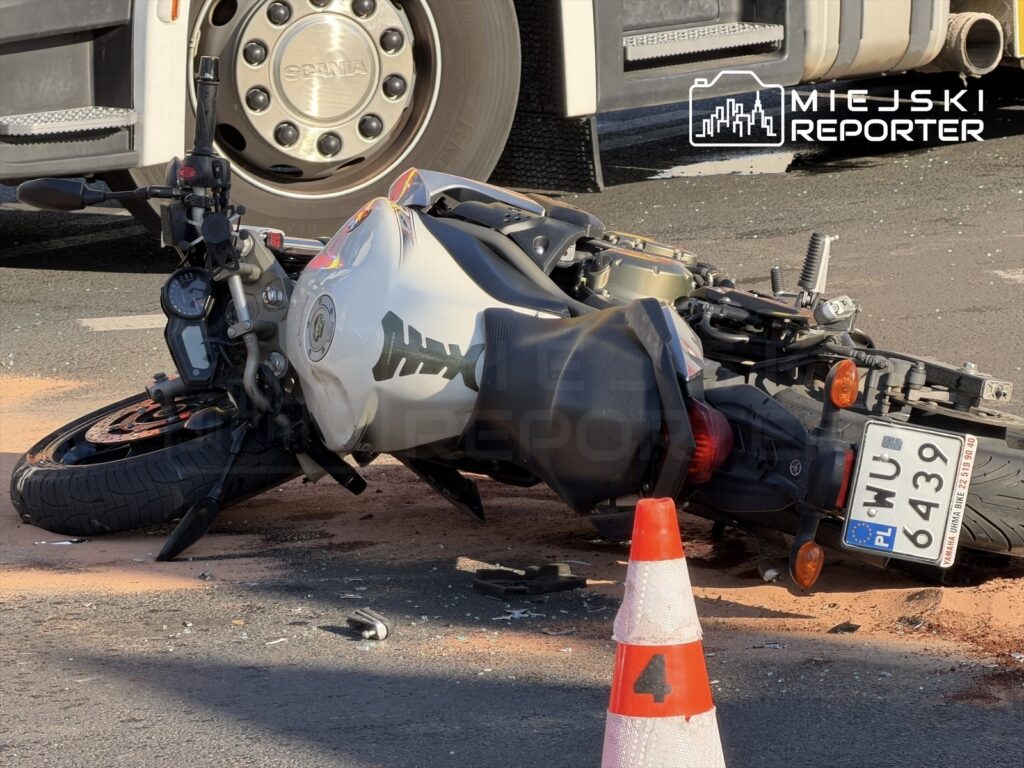 Motocykl leżący na boku na jezdni, obok pomarańczowy proszek, a w tle koło ciężarówki.