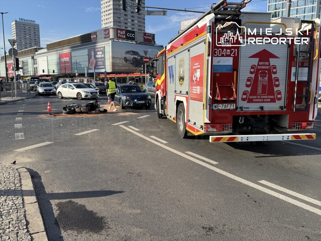 Motocykl leży na jezdni obok wozu strażackiego, a strażak w kamizelce odblaskowej kieruje ruchem w ruchliwym skrzyżowaniu.