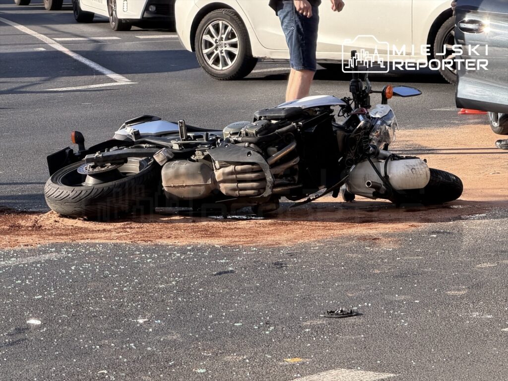 Motocykl leży na boku na drodze, obok niego kilka osób, a w tle widać zaparkowane samochody.