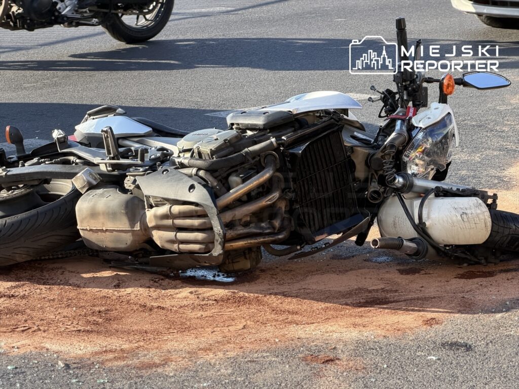 Motocykl leży na boku na jezdni, obok widoczna plama płynu, a w tle przejeżdżające pojazdy.
