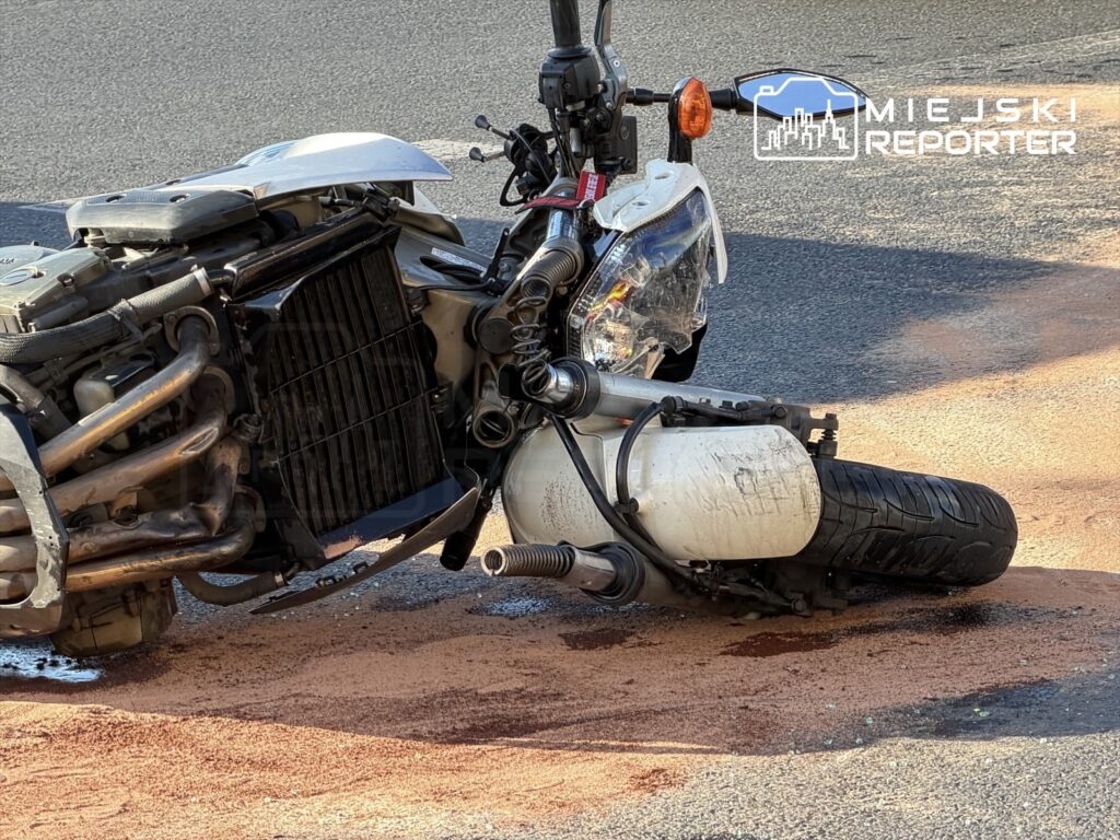 Przewrócony motocykl leży na drodze, wokół niego plama płynów eksploatacyjnych na asfaltowej nawierzchni.