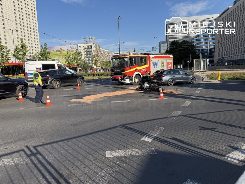 Mężczyzna w kamizelce odblaskowej kieruje ruchem drogowym obok miejsca wypadku, gdzie stoi pojazd straży pożarnej.