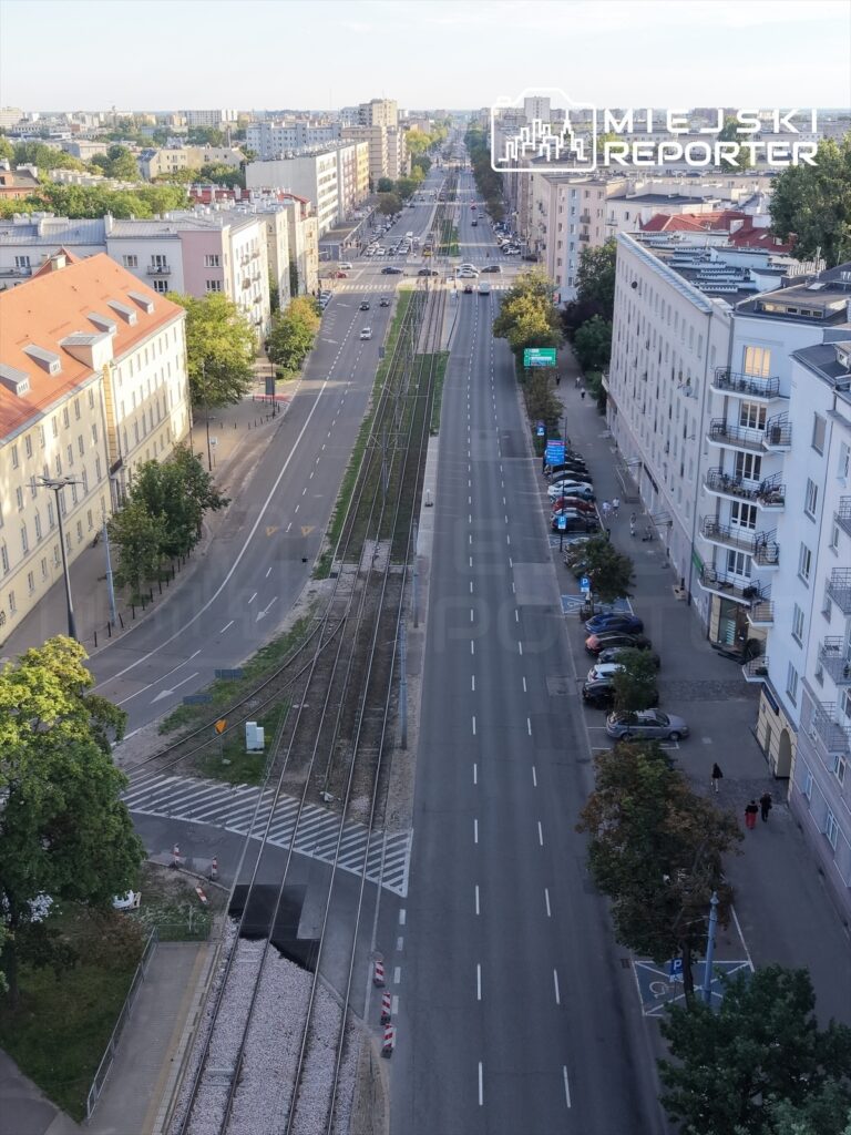 Widok na szeroką ulicę z torami tramwajowymi pośrodku, otoczoną budynkami mieszkalnymi i parkowanymi samochodami.
